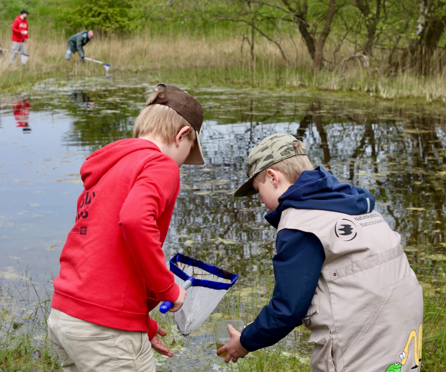 Bilden visar två barn som står vid kanten av en vattensamling, möjligen en sjö eller damm. Det barnet till vänster bär en röd tröja och en keps, och håller i ett litet håvliknande redskap med blått handtag. Det barnet till höger har på sig en beige jacka med en bild av en groda och en ryggsäck. Båda barnen ser ut att vara fokuserade på något i vattnet, kanske de letar efter små vattendjur eller växter att undersöka. I bakgrunden syns en annan person som också verkar vara engagerad i en aktivitet vid vattnet, möjligen också med en håv. Området runt dem är grönt och naturligt med träd och buskar. Det ser ut som en utomhusaktivitet eller utflykt i naturen.